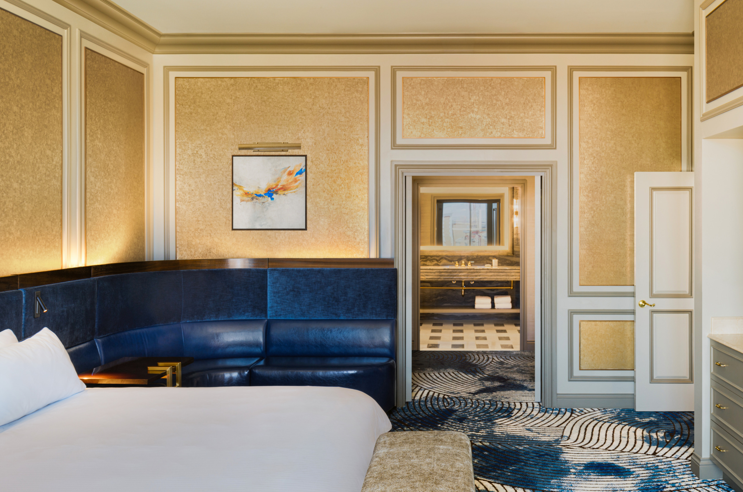Venetian Tower Suite bedroom with curved navy leather headboard, gold textured wall panels, abstract artwork, and a view through to the marble bathroom with black-and-white checkerboard floor.