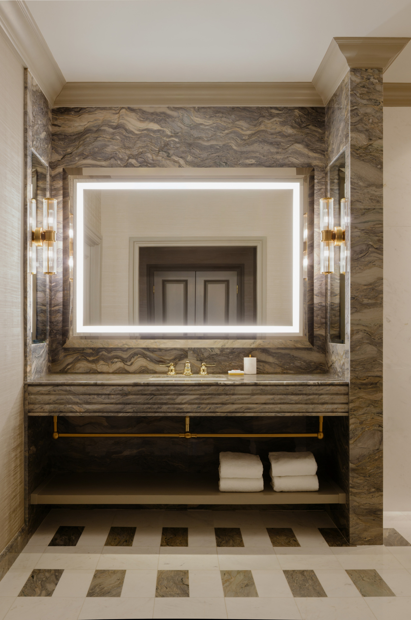 Venetian Tower Suite bathroom vanity with backlit LED mirror, gold fixtures, sconce lighting, waterfall marble walls, and folded white towels on an open shelf.
