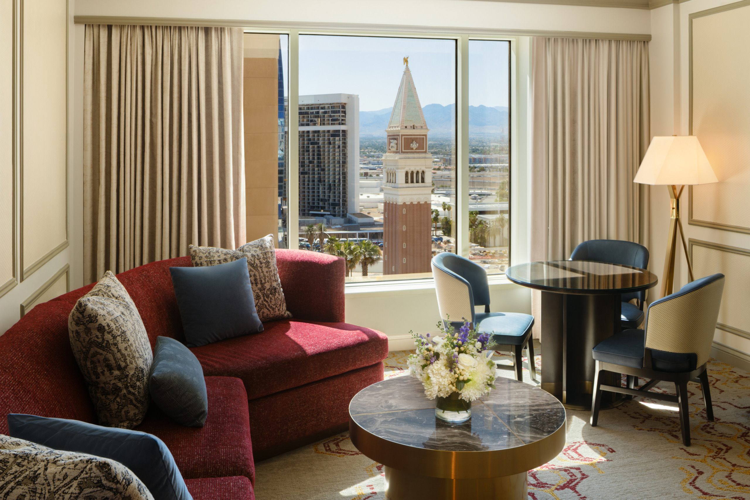 Venetian Tower Suite living room with red patterned sofa, navy accent chairs, marble coffee table, and round dining table, framed by floor-to-ceiling windows showing the Campanile tower and Las Vegas skyline.