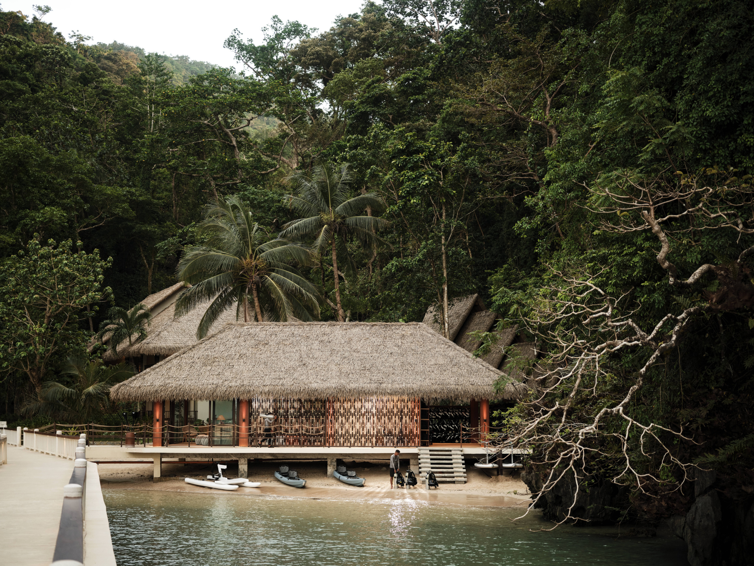 Thatched-roof marine activity center and dive shop at Lagen Island Resort, El Nido, with kayaks and dive gear on the beach below, surrounded by rainforest.