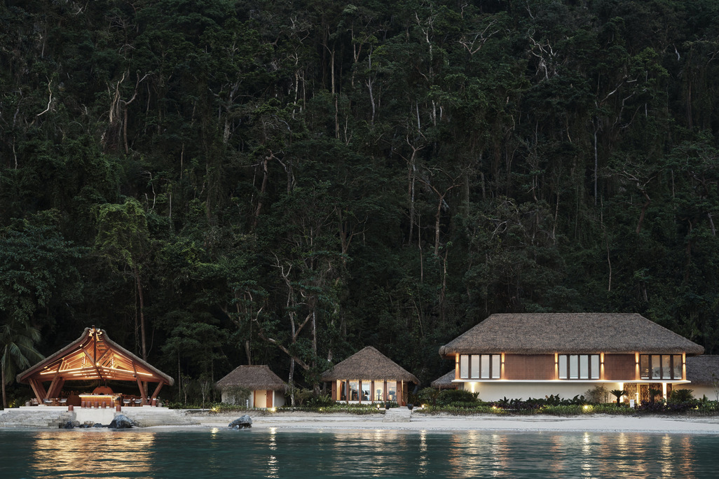 Waterfront view of Lagen Island Resort at dusk, showing timber-frame pavilion on left with warm interior lighting, residential suites with thatched roofs in centre and right, dense tropical forest canopy rising behind, clear lagoon water in foreground.
