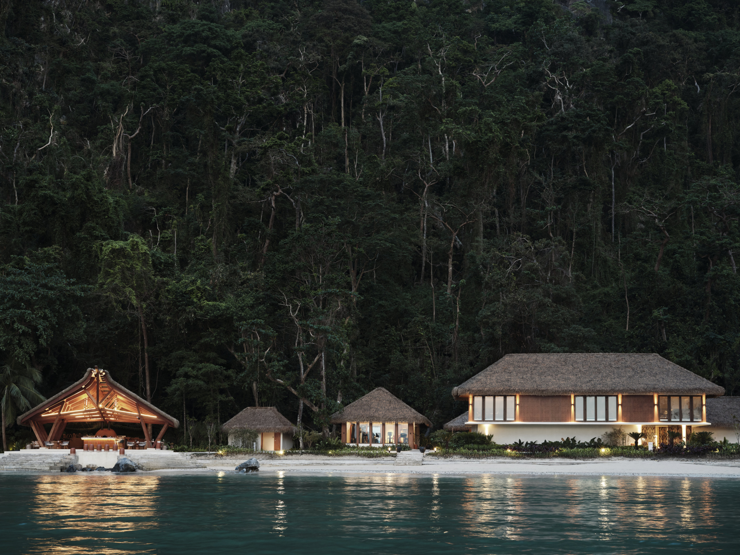 Waterfront view of Lagen Island Resort at dusk, showing timber-frame pavilion on left with warm interior lighting, residential suites with thatched roofs in centre and right, dense tropical forest canopy rising behind, clear lagoon water in foreground.