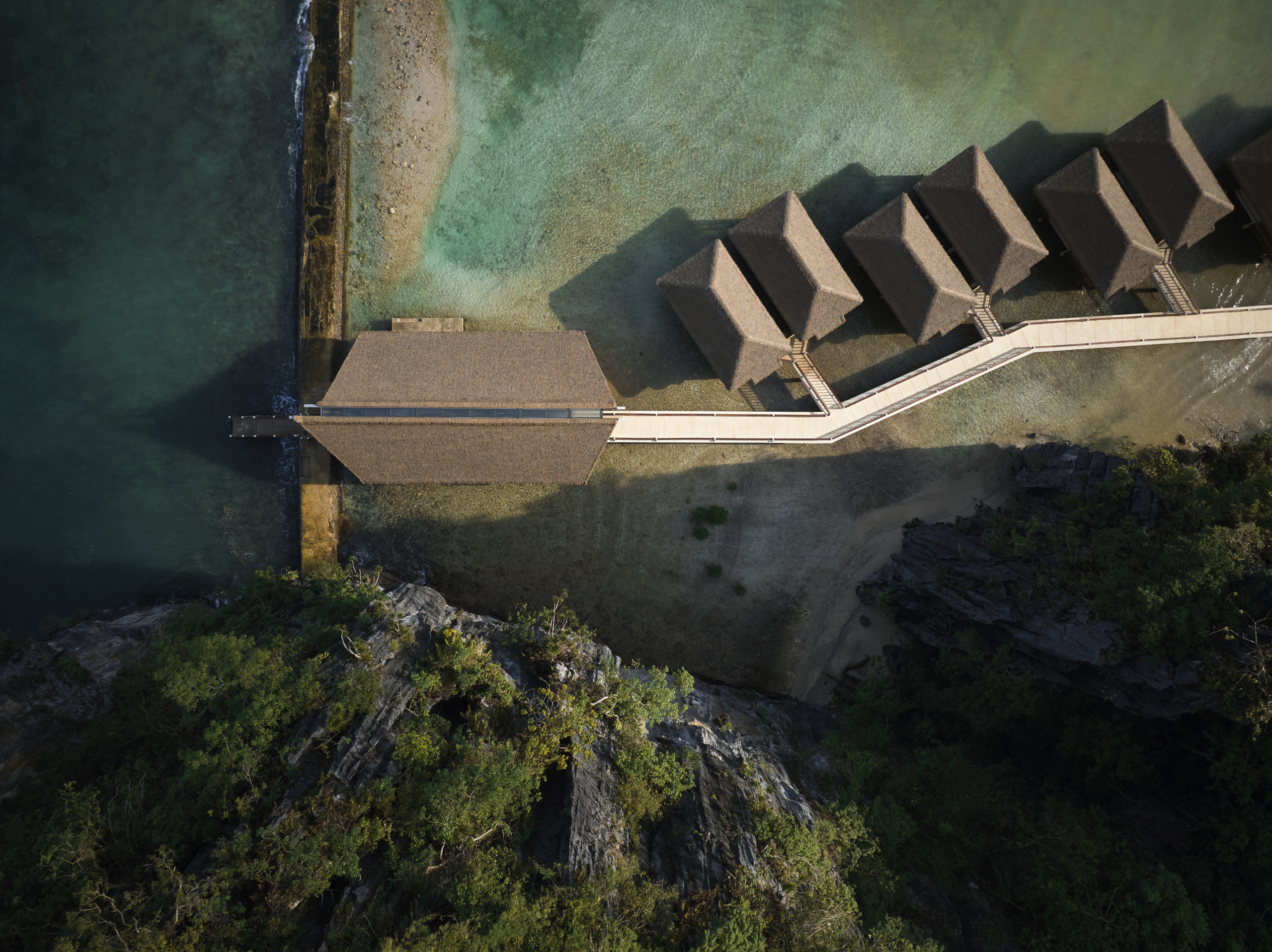 Low-angle view of a traditional thatched overwater pavilion with white railings, set against massive limestone karst formations bathed in warm sunlight at Lagen Island Resort, Palawan.