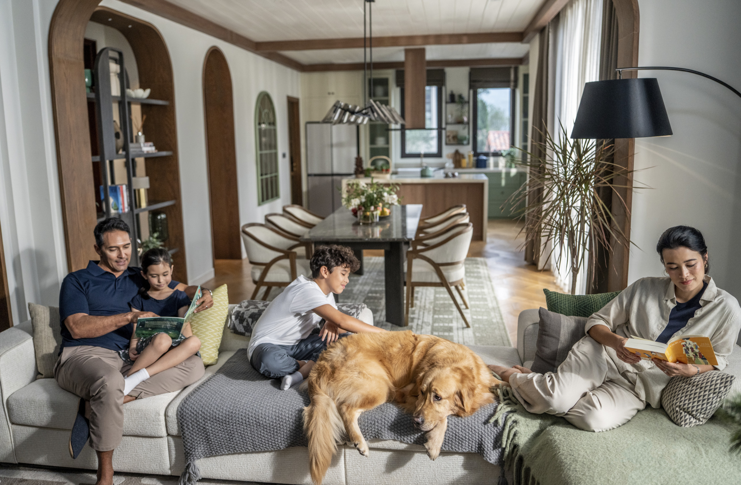 Lifestyle photo of a family relaxing together with a golden retriever in the open-plan living and dining area of a Thanh Xuan Valley residence, featuring warm timber ceiling beams, arched doorways, and a kitchen with sage-green cabinetry in the background.