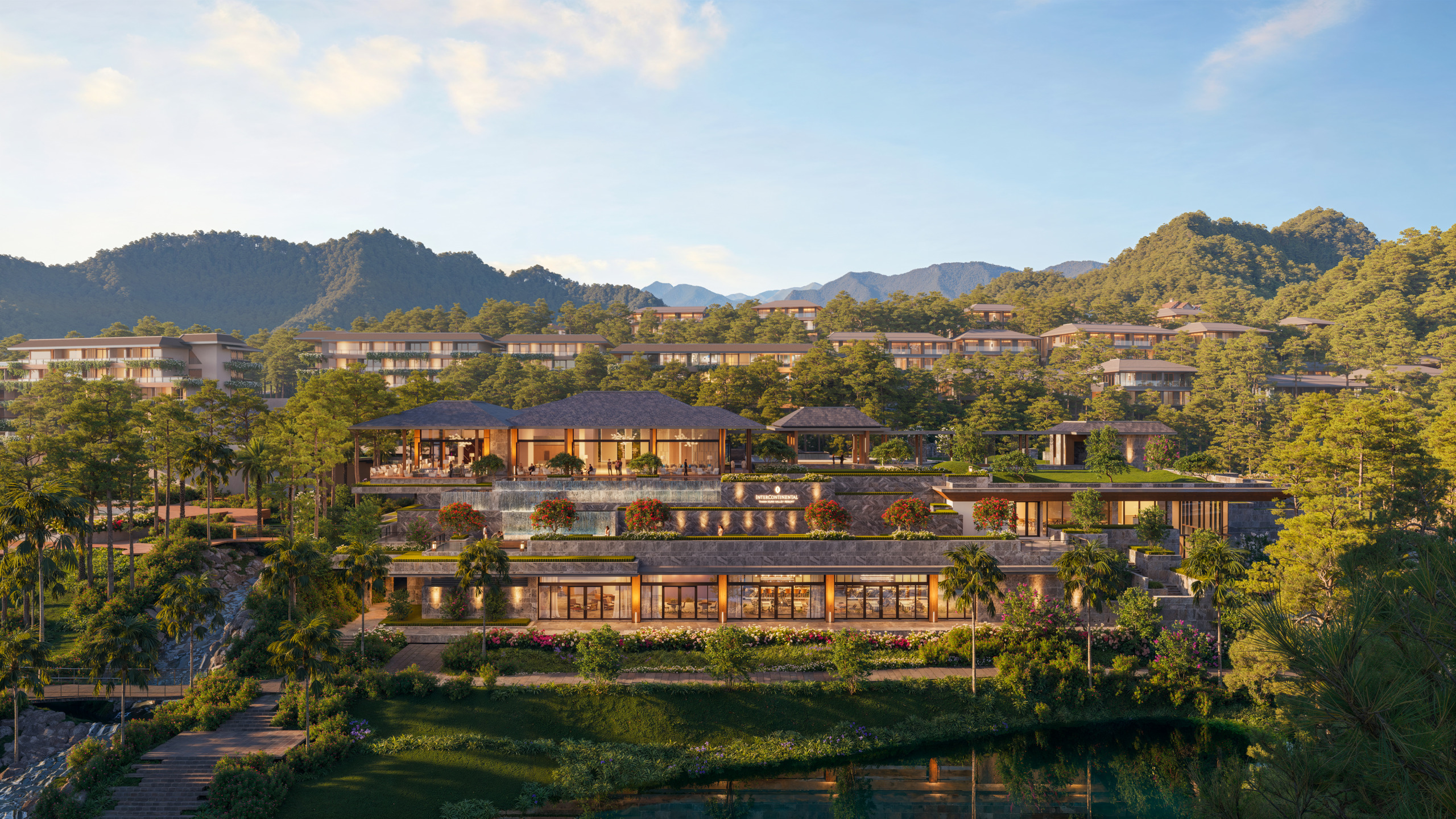 Daytime aerial render of the InterContinental Thanh Xuan Valley Resort, showing tiered stone-and-timber pavilion buildings with pitched roofs terraced down a wooded hillside toward a lake, with residential blocks visible on the slopes behind.