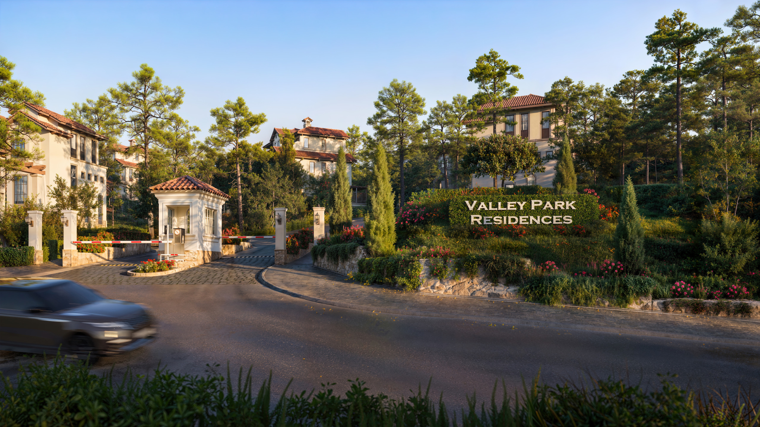Daytime render of the gated entrance to Valley Park Residences at Thanh Xuan Valley, showing a Mediterranean-style guardhouse, a boom barrier over a tree-lined drive, and a hedge sign spelling out the residence name, with a car passing on the road in the foreground.