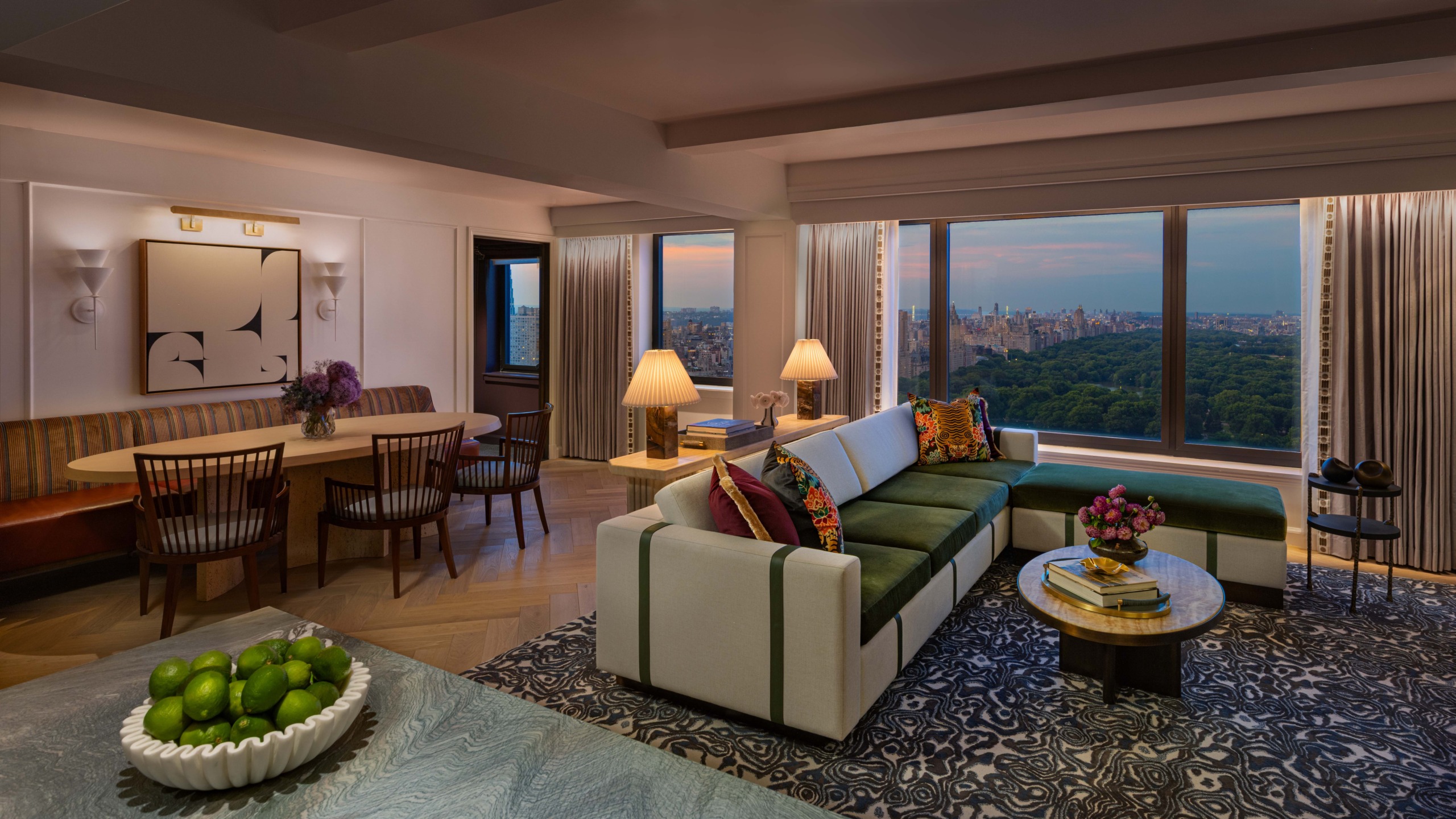 Promenade Suite living and dining area at dusk with green sectional sofa, dining table, and floor-to-ceiling views of Central Park, JW Marriott Essex House.