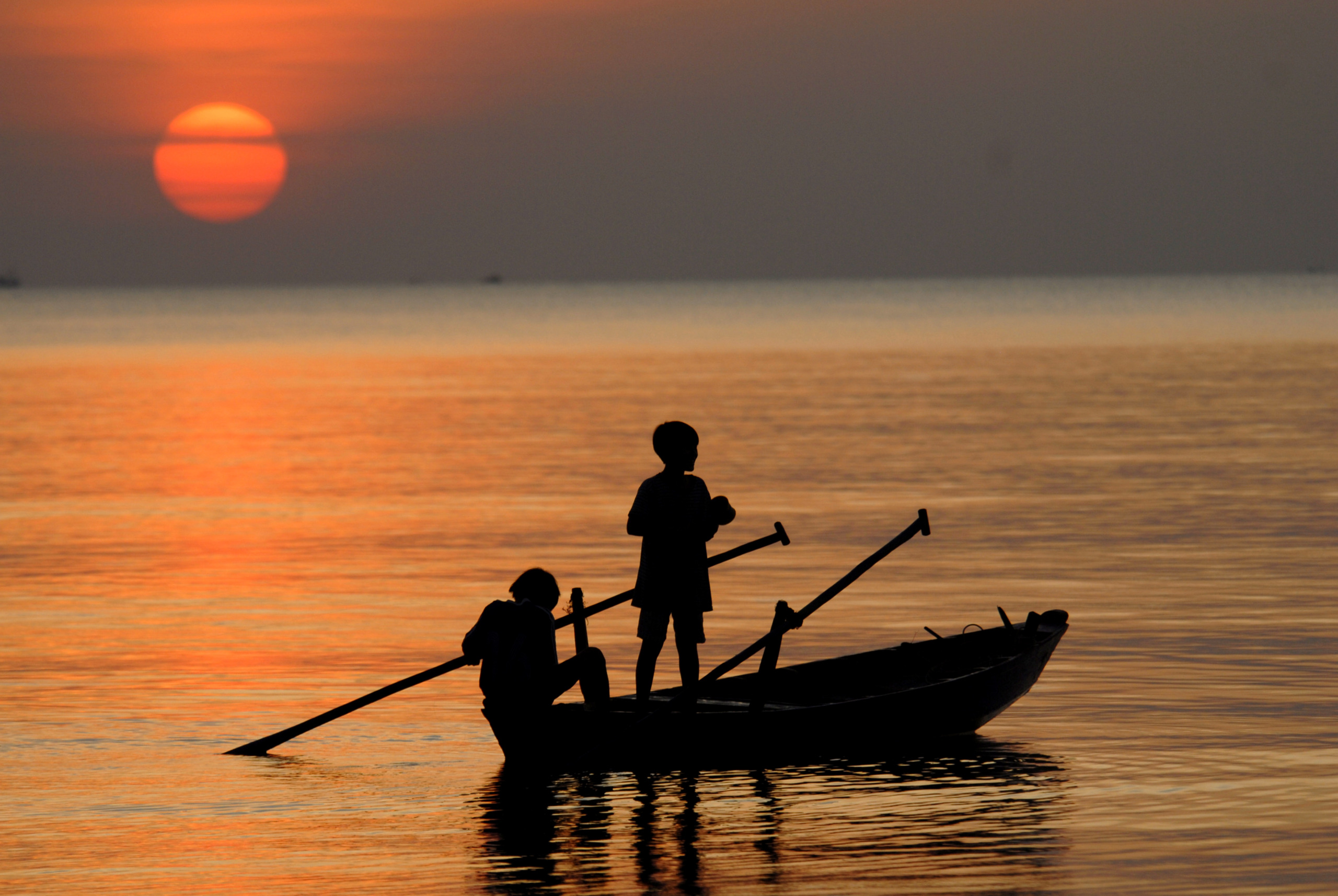 Silhouette of two people rowing a small wooden boat at sunset in Phu Quoc, Vietnam, with the sun low over calm water casting an orange glow across the sea.