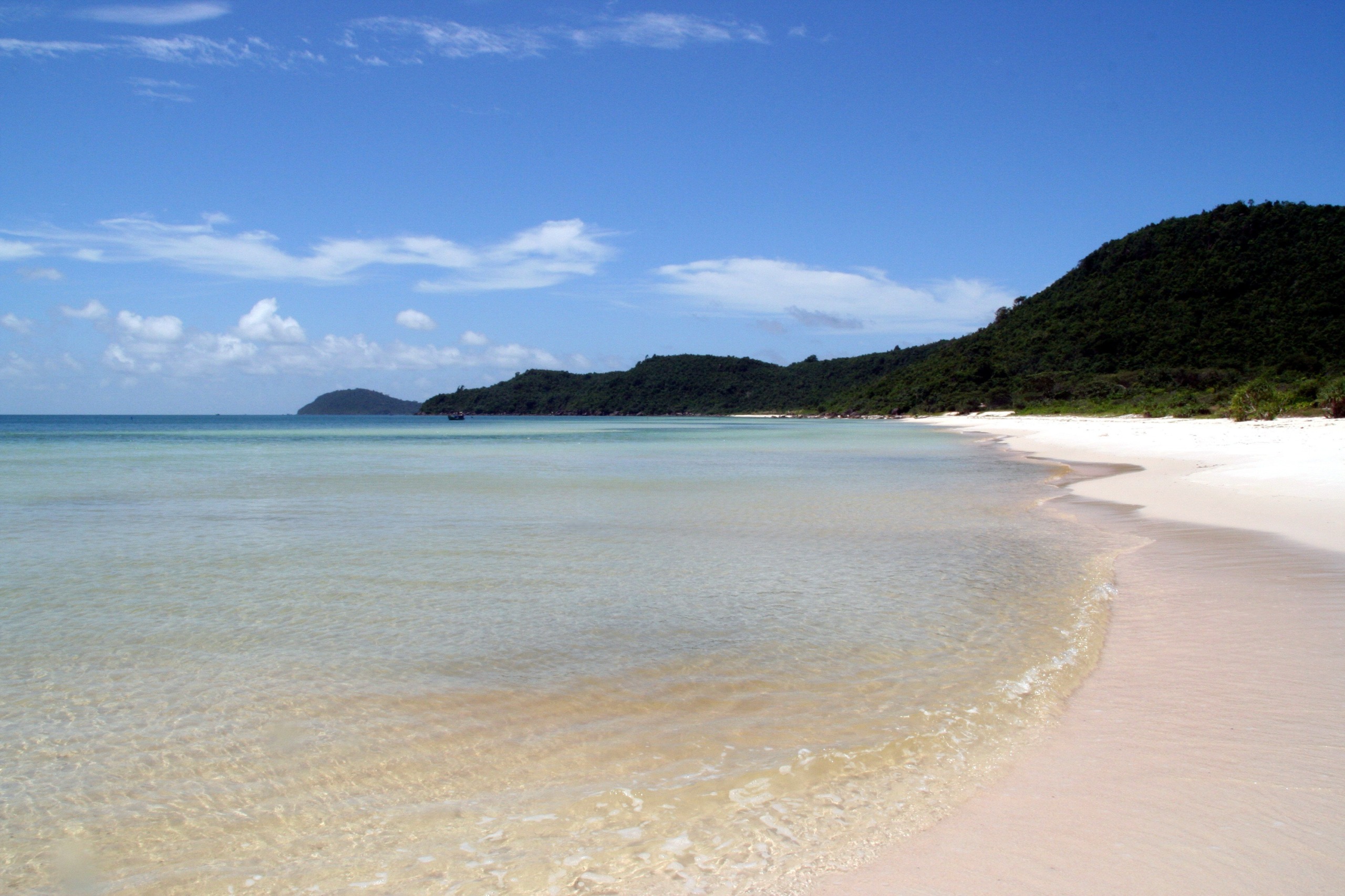 White sand beach and clear turquoise water along the coast of Phu Quoc, Vietnam, with forested hills stretching along the shoreline under a blue sky.