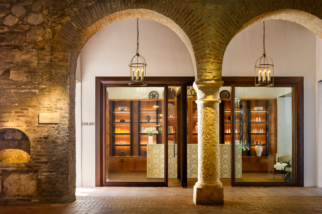 Umari Spa boutique entrance at Four Seasons Resort Cartagena, framed by a stone archway and column, with a wood-and-glass storefront displaying lit retail shelves, ceramics, and hanging lantern fixtures.