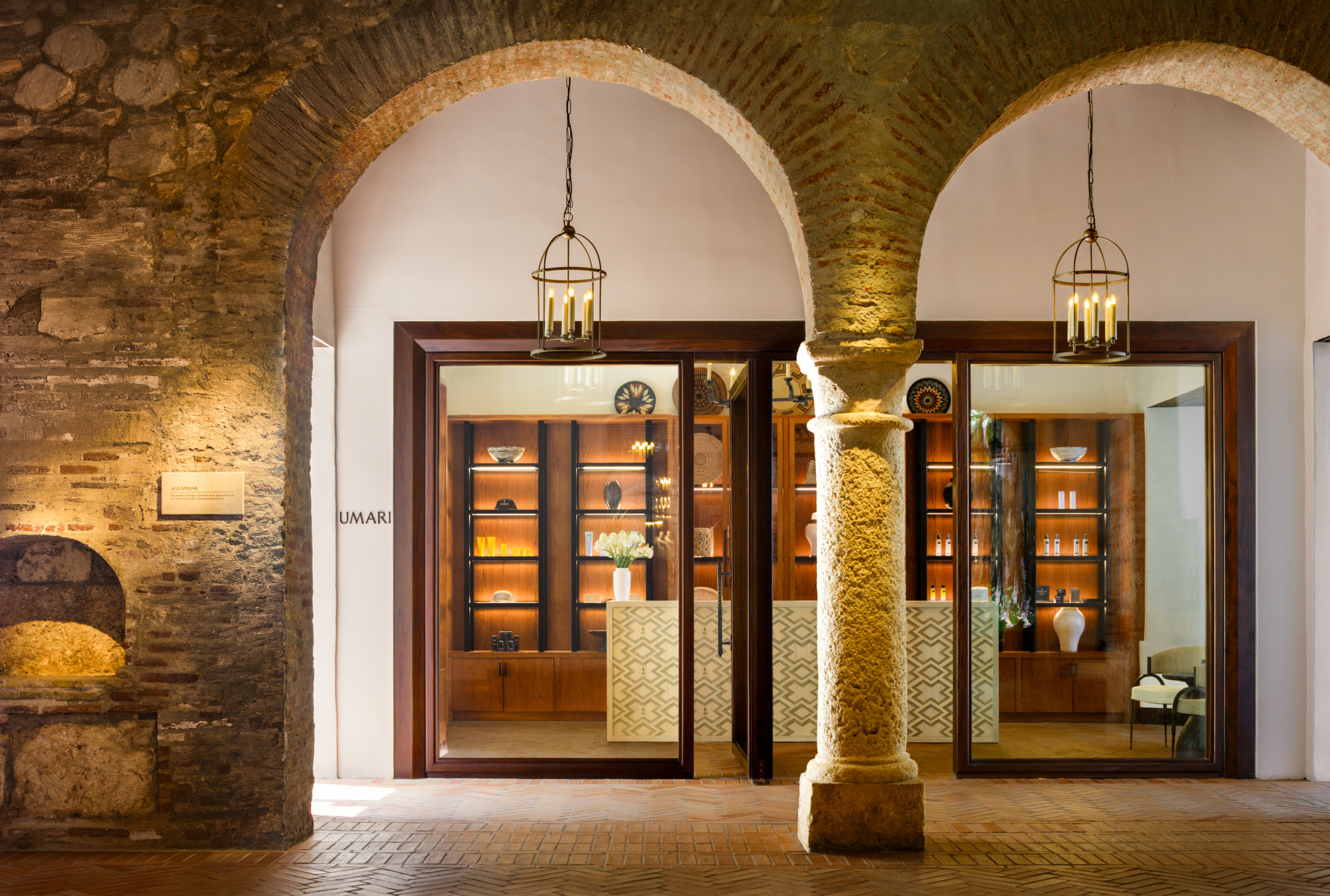 Umari Spa boutique entrance at Four Seasons Resort Cartagena, framed by a stone archway and column, with a wood-and-glass storefront displaying lit retail shelves, ceramics, and hanging lantern fixtures.