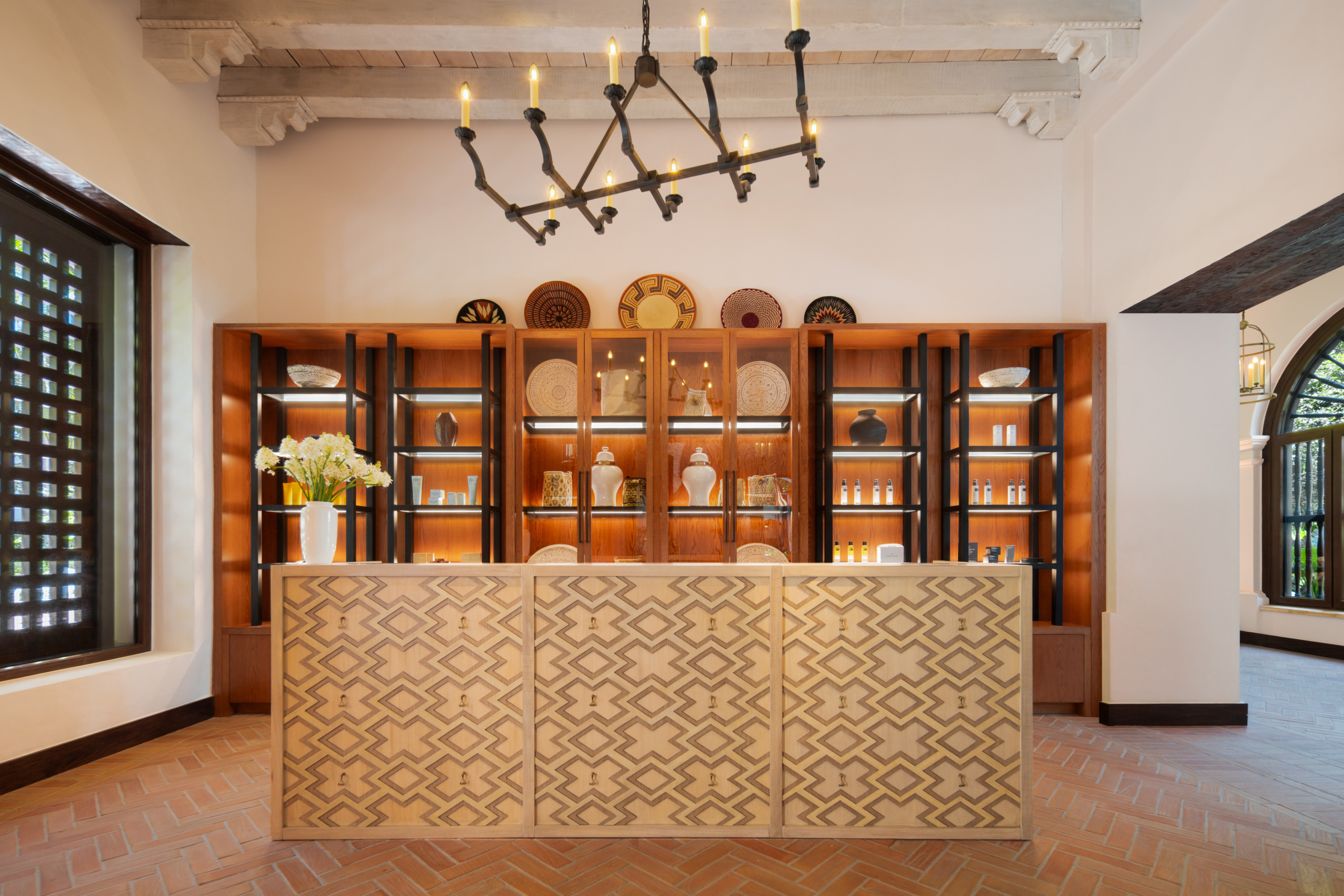 Umari Spa reception desk at Four Seasons Cartagena, featuring a geometric wood-inlay counter, illuminated shelving with ceramics and skincare products, and a wrought-iron candle chandelier overhead.