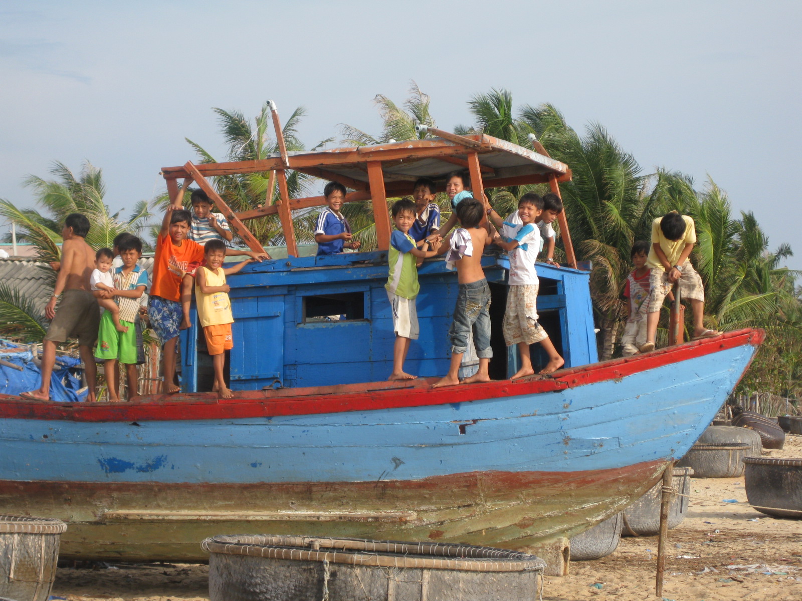 Group of children playing and posing on a weathered blue fishing boat beached on the sand in Phu Quoc, Vietnam, with palm trees and woven coracle boats in the background.