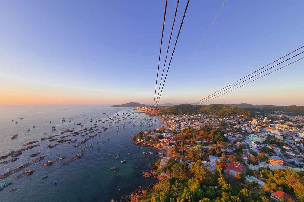 Aerial view from a cable car over Phu Quoc, Vietnam, showing a dense cluster of fishing boats anchored in a turquoise bay, a coastal fishing village with colorful rooftops and palm trees, and forested hills stretching along the coastline at golden hour.