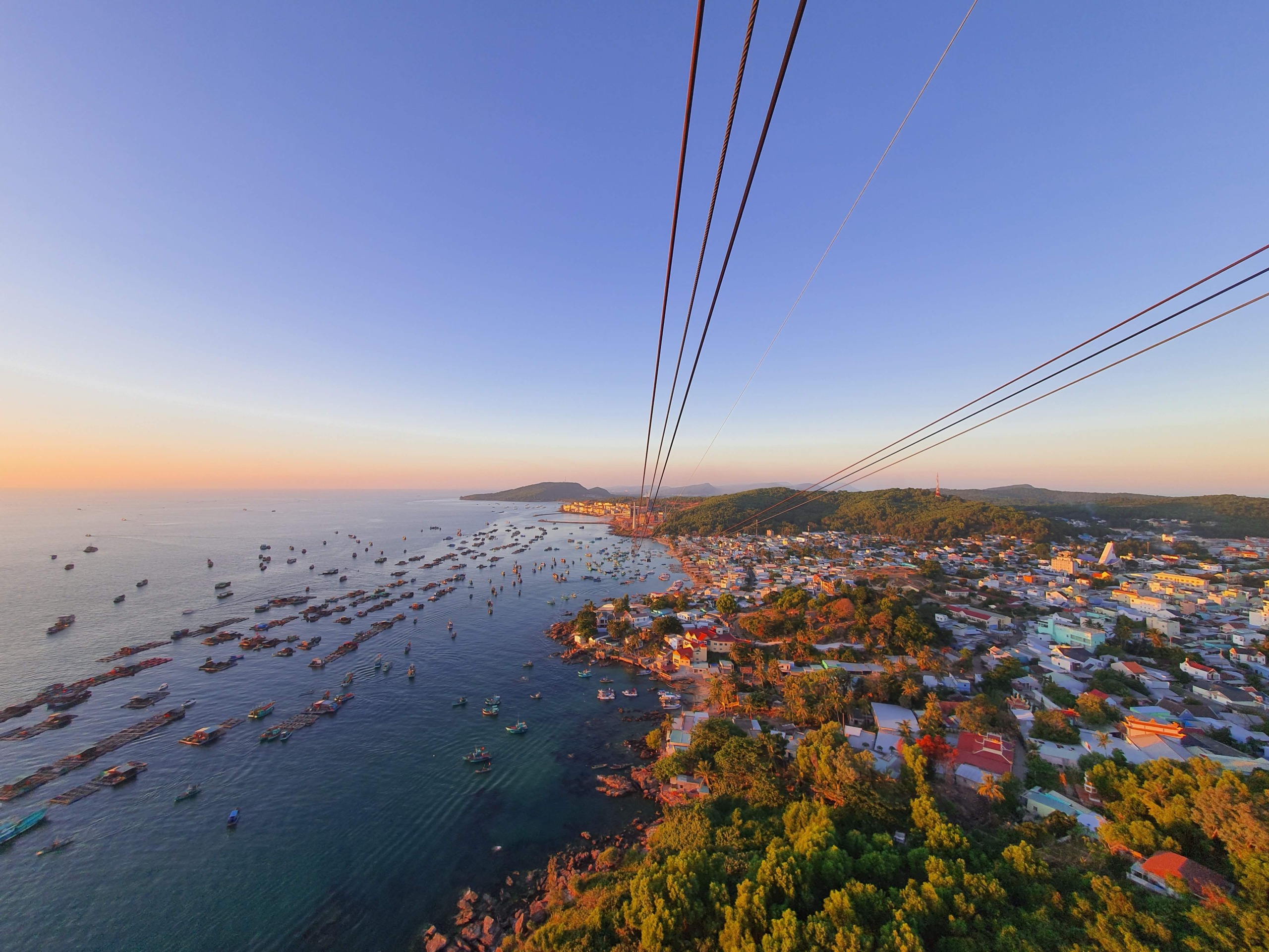 Aerial view from a cable car over Phu Quoc, Vietnam, showing a dense cluster of fishing boats anchored in a turquoise bay, a coastal fishing village with colorful rooftops and palm trees, and forested hills stretching along the coastline at golden hour.