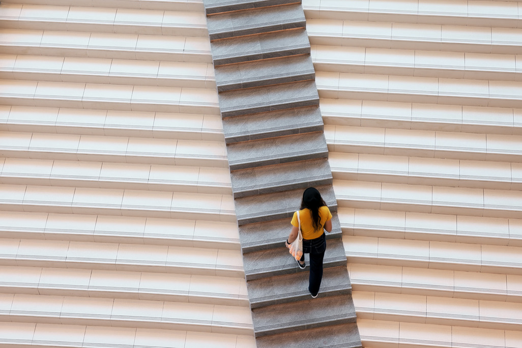 Emerging designer climbing central staircase between two lighter flights of steps, carrying architecture portfolio, viewed from above