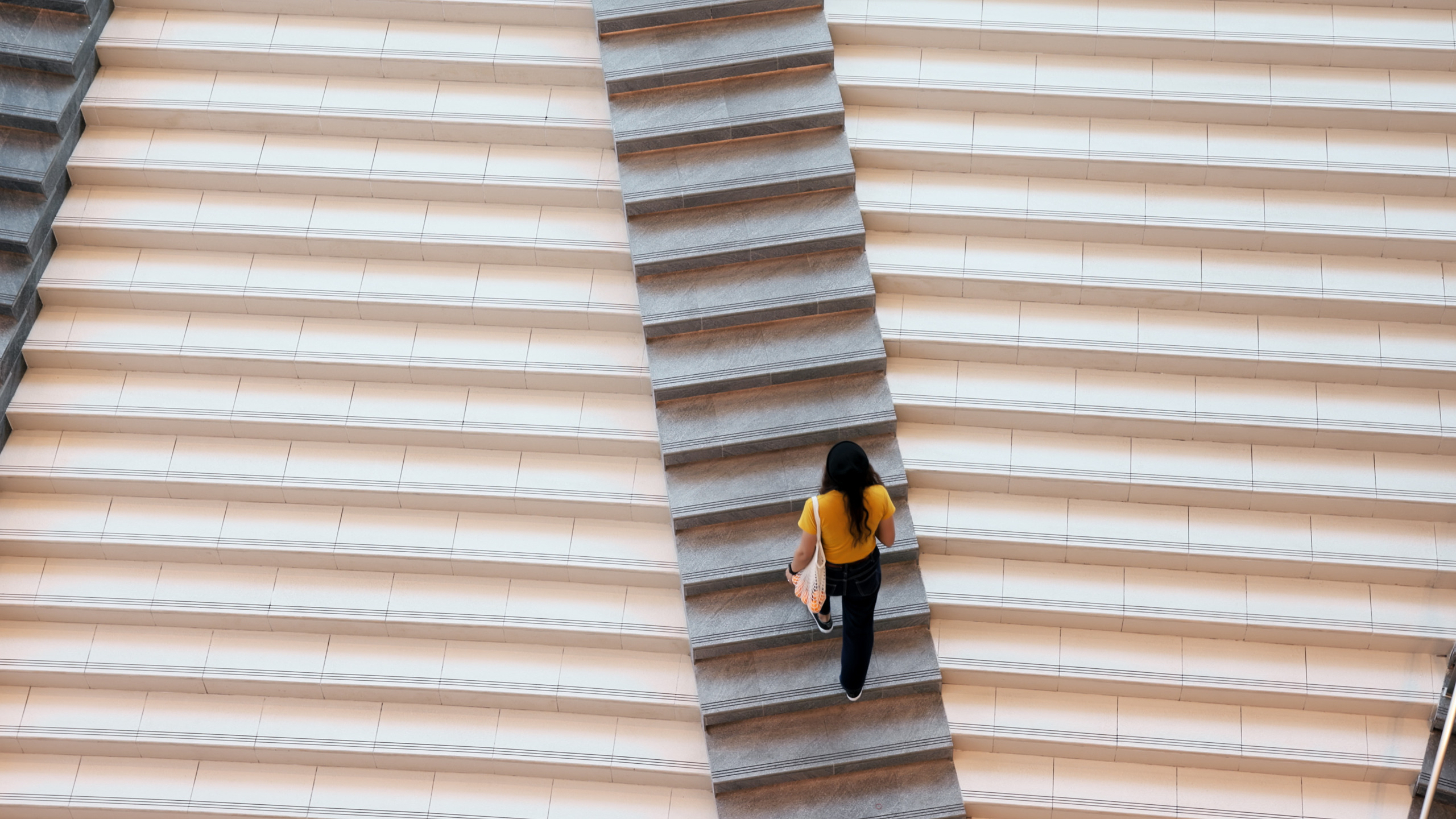 Emerging designer climbing central staircase between two lighter flights of steps, carrying architecture portfolio, viewed from above