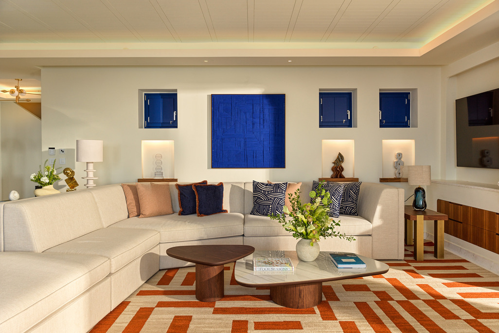 Living room at Four Seasons Mykonos with a curved cream sofa, cobalt blue artwork, walnut tables and an orange geometric rug.