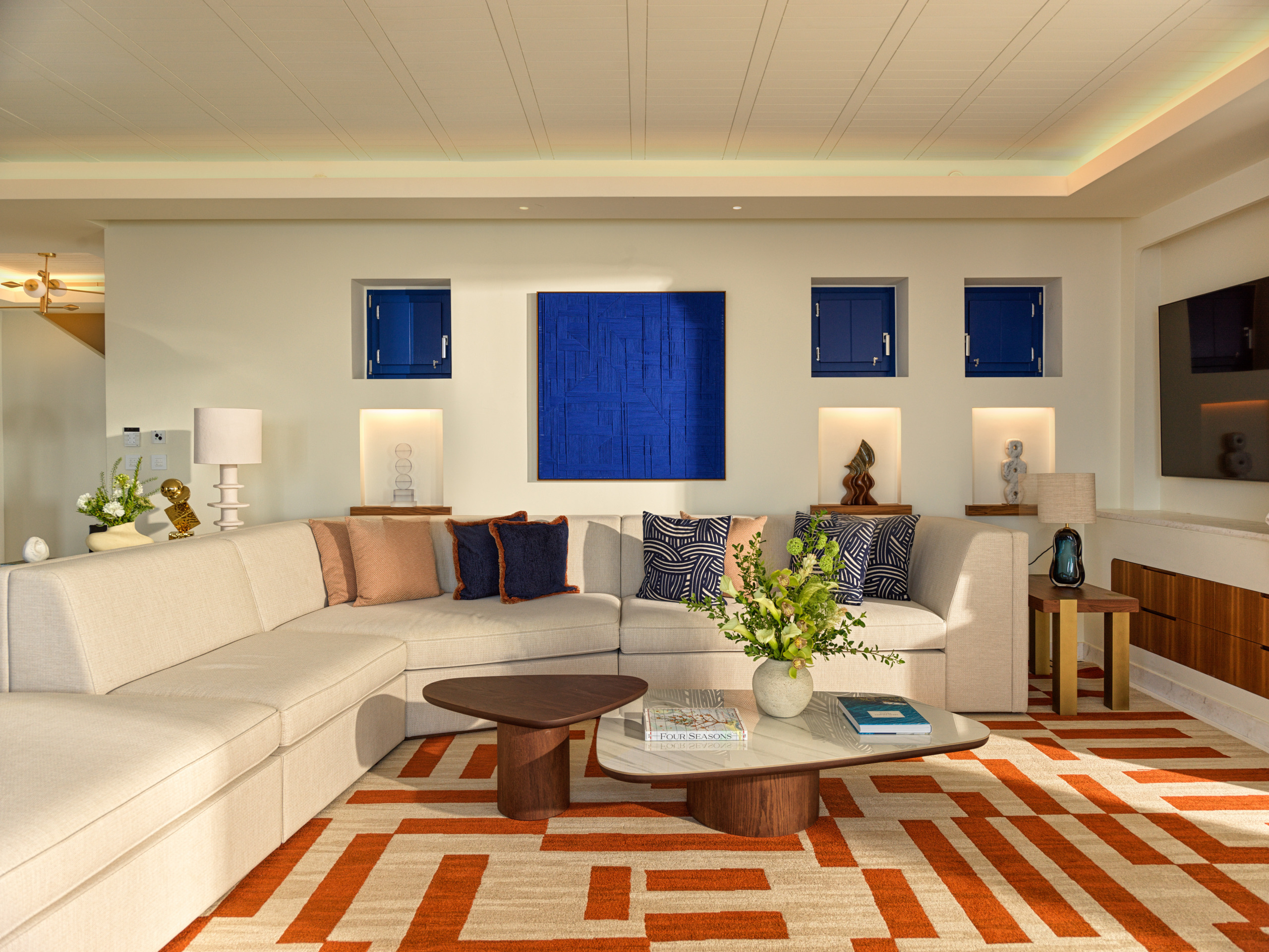 Living room at Four Seasons Mykonos with a curved cream sofa, cobalt blue artwork, walnut tables and an orange geometric rug.