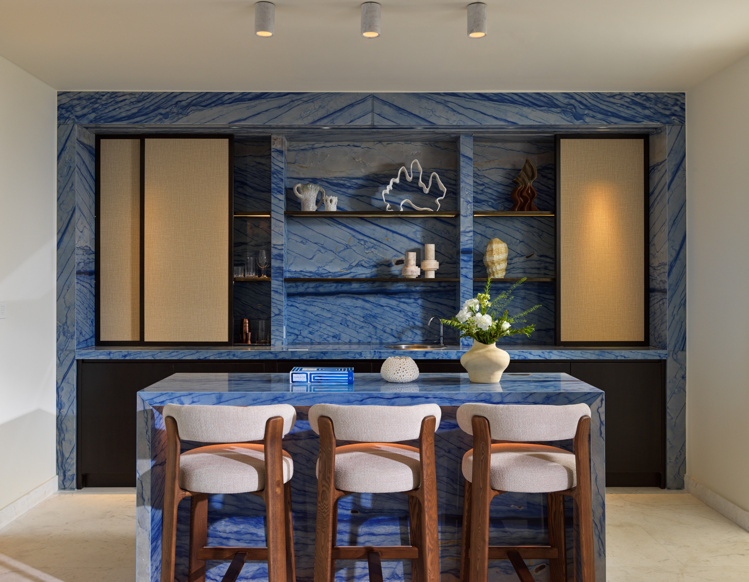 Suite bar at Four Seasons Mykonos clad in blue-veined stone, with a waterfall island, three counter stools and open shelves.
