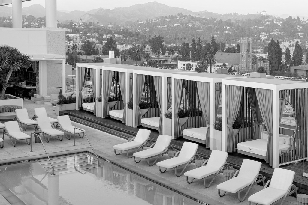 W Hollywood rooftop pool with white sun loungers, draped cabanas, and hillside views beyond.