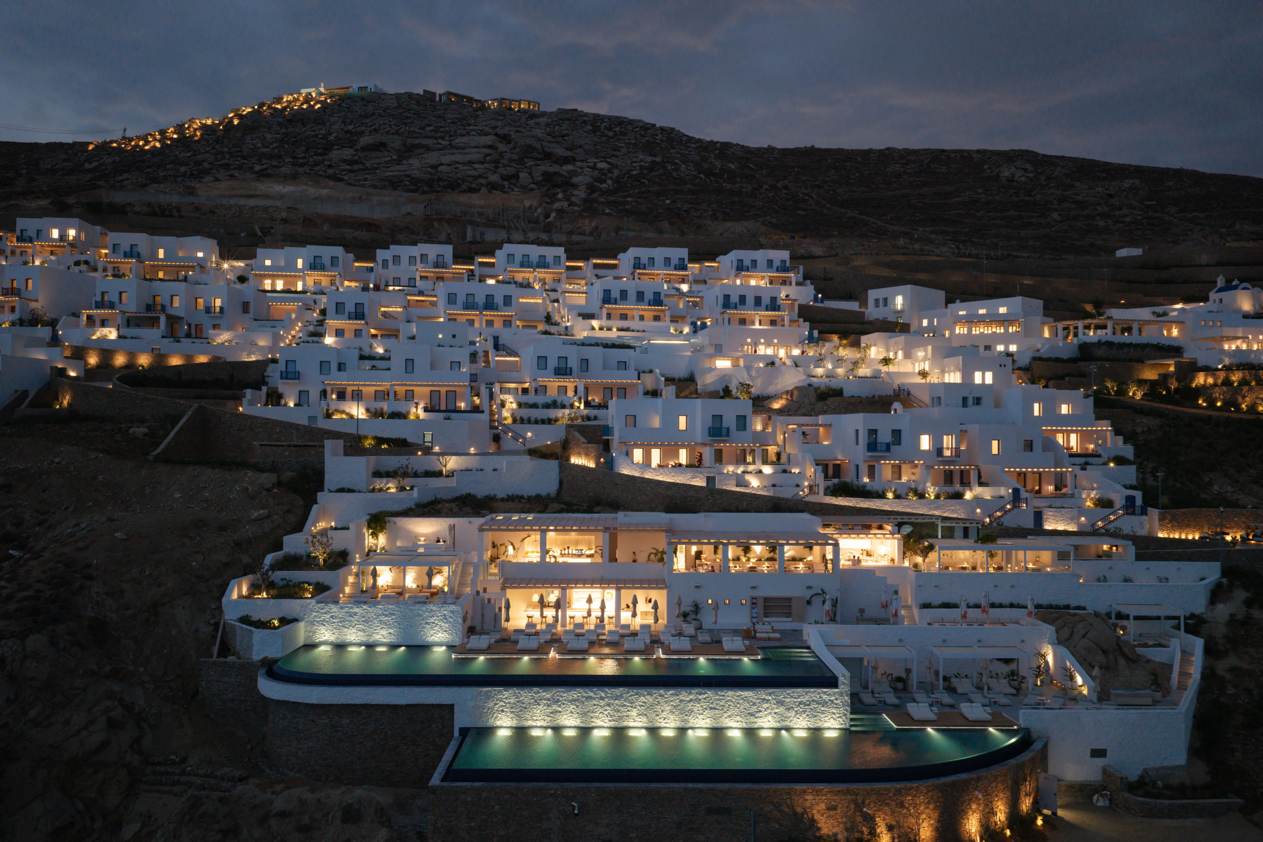 Aerial dusk view of Four Seasons Mykonos: whitewashed buildings terraced down a hillside above two tiered infinity pools.